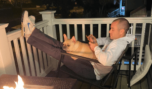 Male sitting on patio with his dog, scrolling on his phone next to a fire.