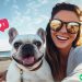 A fun-loving brunette female holding her Iphone and taking a selfie with her cream colored french bulldog on the boardwalk at the beach. There is an Instagram heart icon above the dog's face.