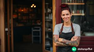 a quirky looking woman with dark red hair and tattoos standing outside of her bakery with her arms crossed, smiling at the camera.