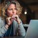 A mother holding a pen to her lips with a pensive look on her face as she looks at her laptop, suspicious of a scam.