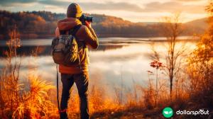 A man with a backpack and a camera taking a picture of a beautiful lake during the fall season.