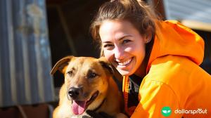 A woman wearing an orange jacket kneeling down next to a dog. Both are smiling at the camera.