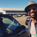 A man wearing a hat and sunglasses smiling at the camera. He is standing next to his car that he Ubers with. He is at an airport.