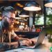 A hipster-looking guy making money from his laptop at a coffee shop.