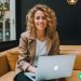 A woman sitting on a leather couch with her Macbook, smiling at the camera. She is a freelance writer.