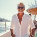 A wealthy older man standing on his yacht in an exotic location with a smile on his face.