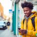 A man in his twenties standing on a city sidewalk as he looks down at his phone with a soft smile on his face.