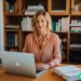 A 38 year old woman proudly sitting in her home office with her laptop out.