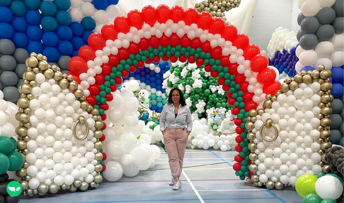 Karin Capellan standing under a large, rainbow-colored balloon arch inside a gymnasium, surrounded by elaborate balloon sculptures.