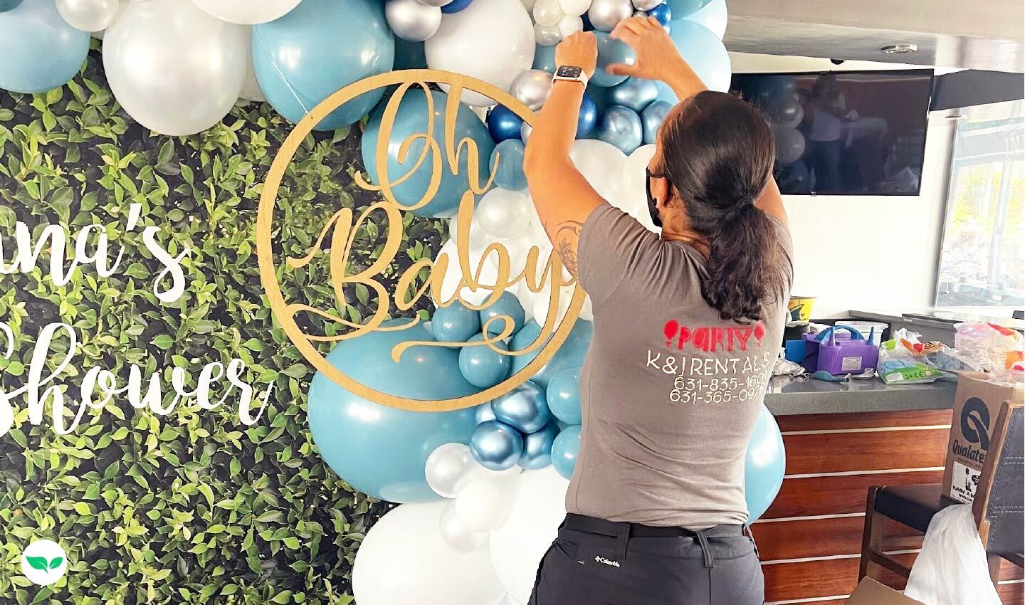 Karin Capellan working on a blue and white balloon installation for a baby shower, adjusting decorations on a lush greenery backdrop.