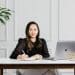 Linda Tran sits at her desk in a sunlit home office, working on a laptop with notebooks, pens, and presentation materials nearby.