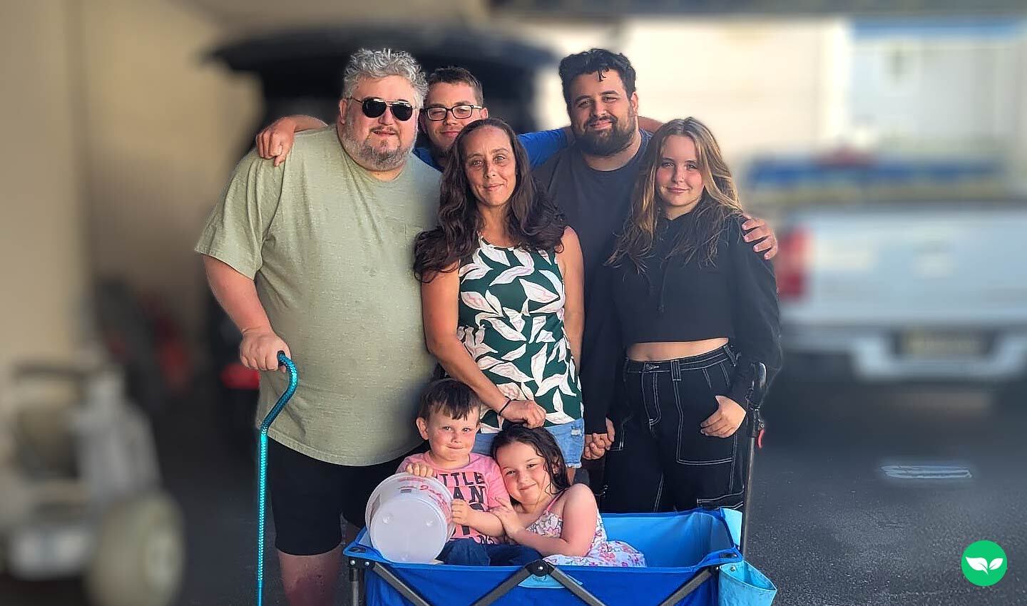 Brian Watkins and his blended family smiling together at the shore, standing around a blue wagon with two young kids sitting inside — a glimpse of the life he rebuilt after losing his home in a fire.