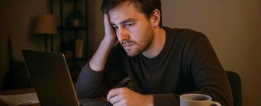 A tired man sits at his desk late at night, staring at his laptop with a cup of coffee nearby — representing the burnout many side hustlers feel after long hours of extra work.