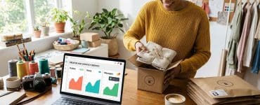 Woman in a bright home studio packaging handmade products for shipment, with a laptop showing order and sales growth charts beside her.